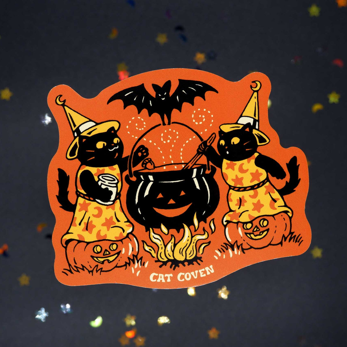 A die-cut sticker featuring a Halloween cat illustration, displayed on a dark gray background with scattered gold and silver confetti. The sticker shows two black cats in orange patterned dresses and hats standing beside a black cauldron with flames, a bat, and jack-o'-lanterns, with the text 'CAT COVEN' at the bottom.