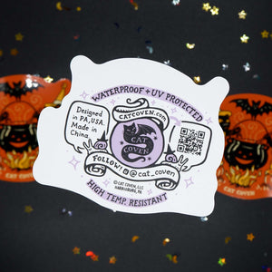 A white die-cut sticker back with purple and black cat-themed graphics and text, shown in front of two orange Halloween decorations on a dark background with scattered gold star confetti.