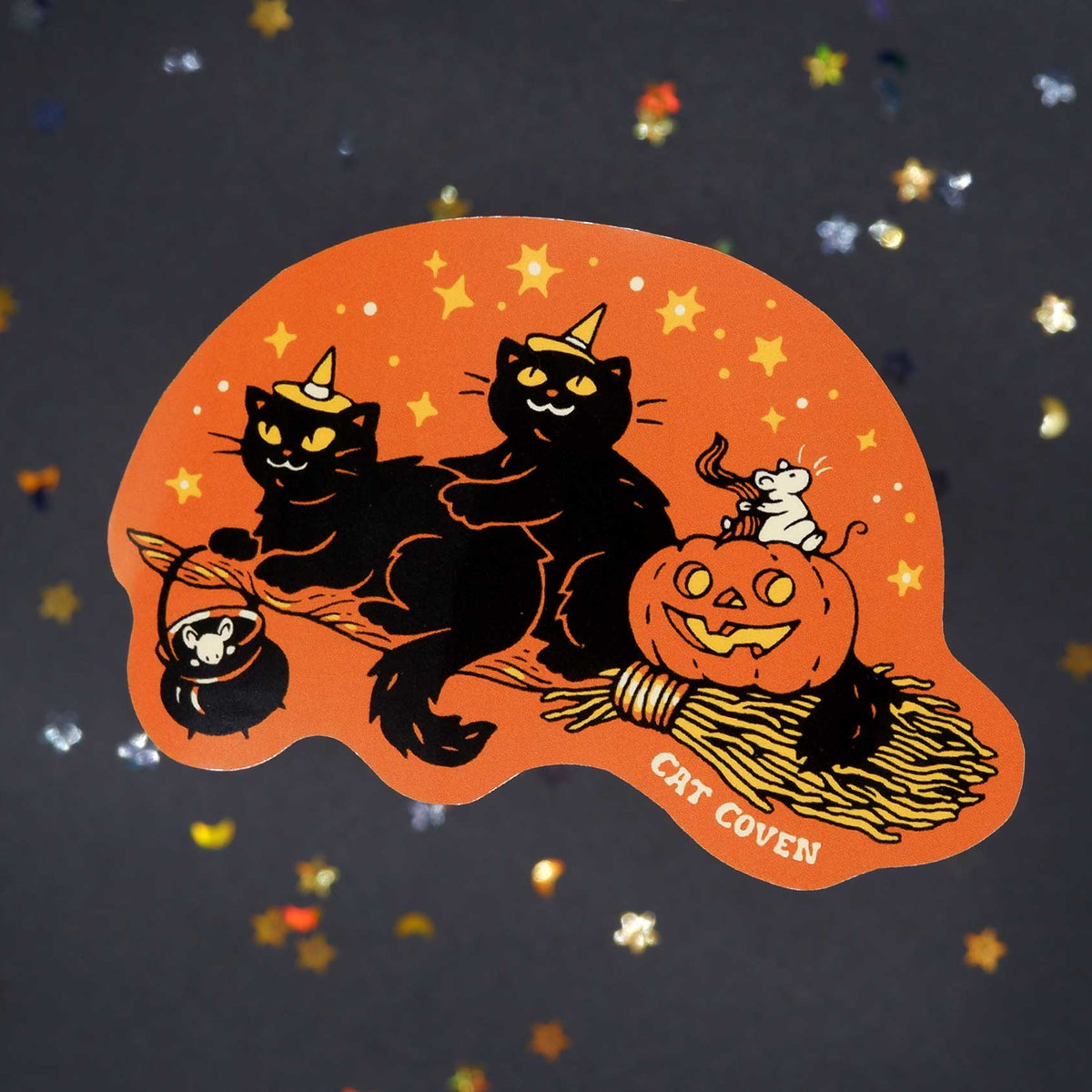 A die-cut sticker featuring a Halloween cat illustration on an orange background, with two black cats, a jack-o'-lantern, two white mice, and the words 'CAT COVEN' on a dark glittery surface.