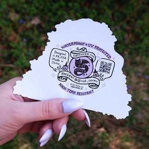 Hand holding a white sticker back  with black text and design against a blurred green background