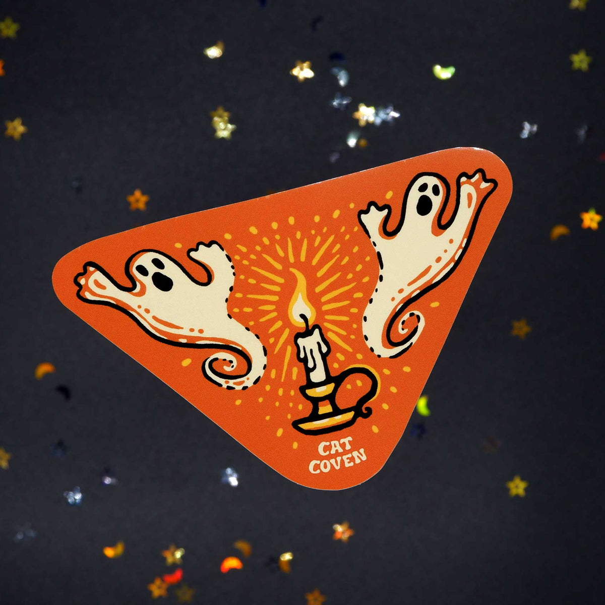 An orange triangular sticker featuring a Halloween design with two white ghosts, a candle, and the text 'CAT COVEN' on a dark background with scattered star confetti.