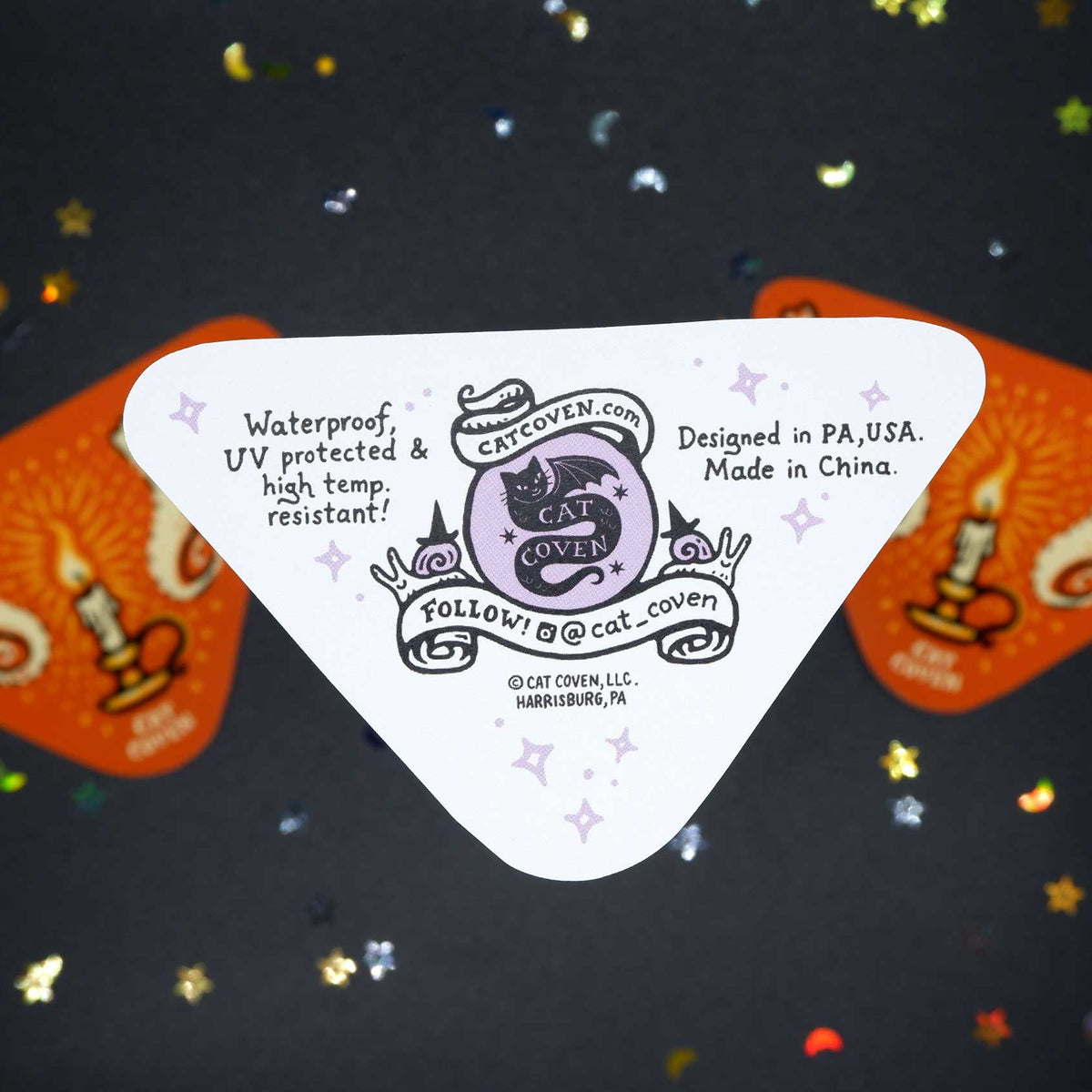 A white triangular sticker back with printed black text and a lavender cat logo reading catcoven.com, placed on top of two orange decorative cards against a dark glittery background.