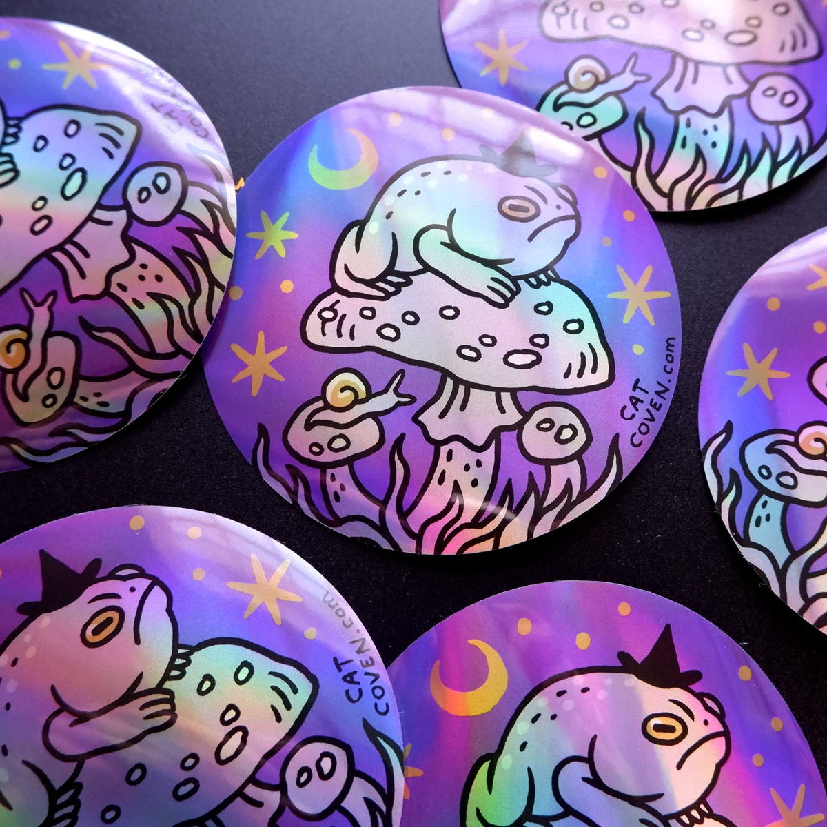 Several round stickers with holographic iridescent finishes, featuring a whimsical illustration of a toad sitting on a mushroom under a crescent moon and stars, displayed on a dark background.