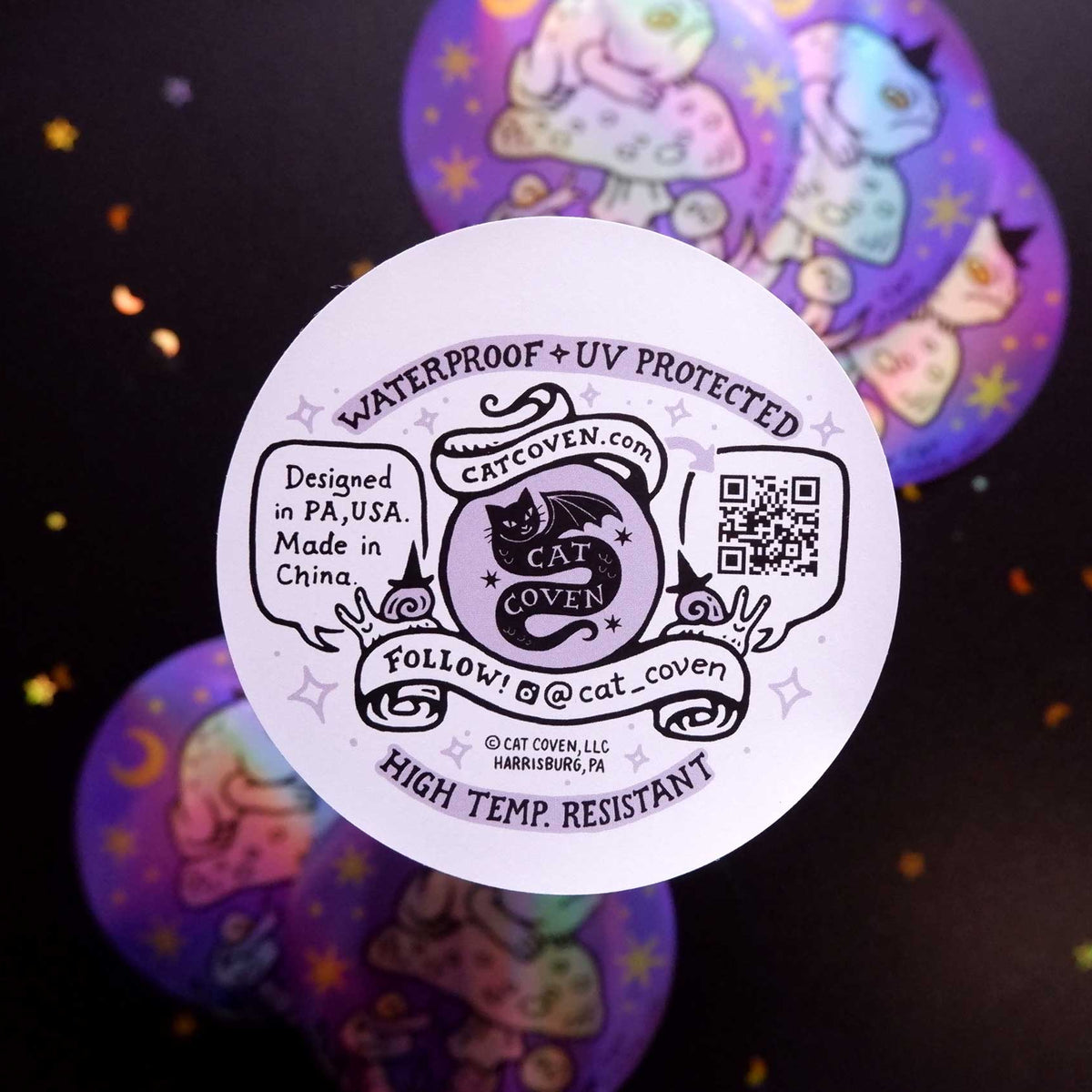 A round white sticker back with black printed text and a cat logo, shown in front of three overlapping purple circular graphics on a dark background with small gold confetti-like specks.