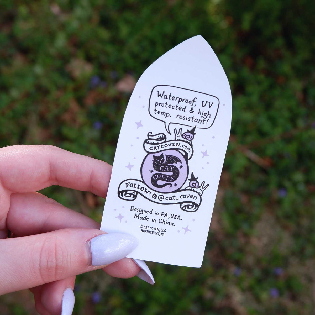 Hand holding a small white sticker back with black text and graphics against a blurred green background
