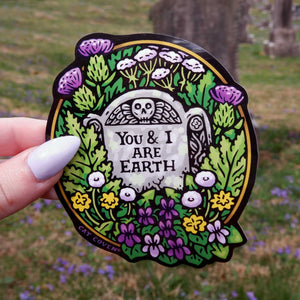 Decorative sticker with 'You & I Are Earth' text held in front of a natural background