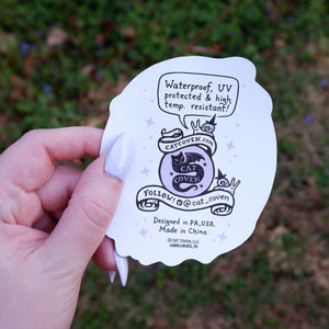 Hand holding a white sticker back with black text and graphics against a blurred green outdoor background