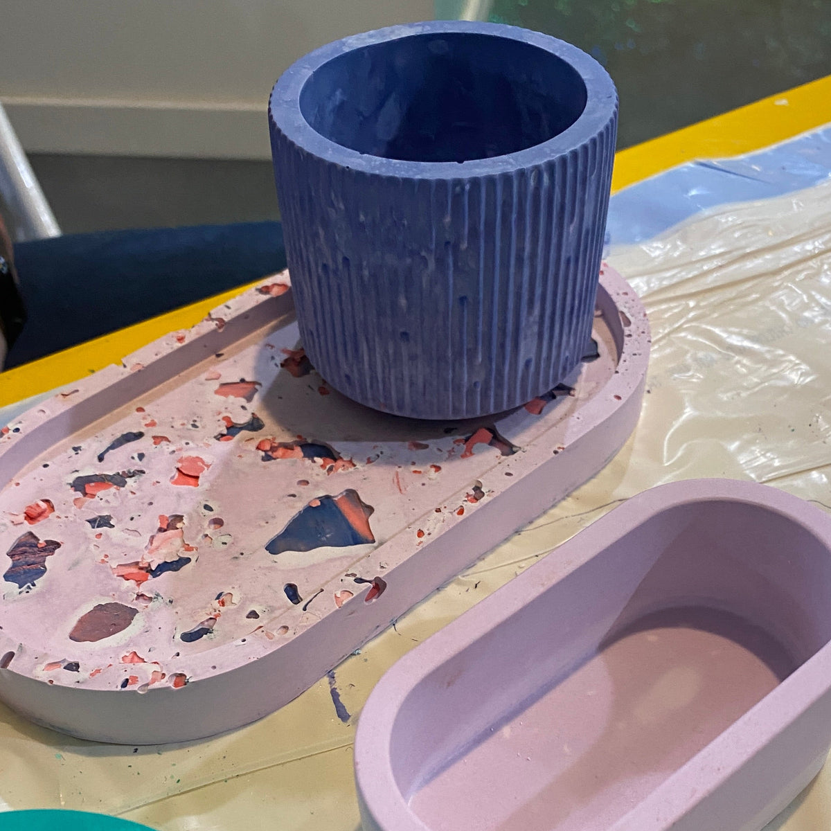Colorful ceramic items including a blue cup, purple oval container, and turquoise lid on a textured surface.