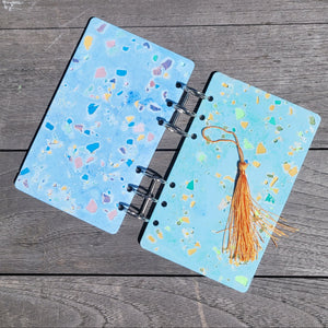 Open notebook with terrazzo design and a tassel on a wooden surface. 