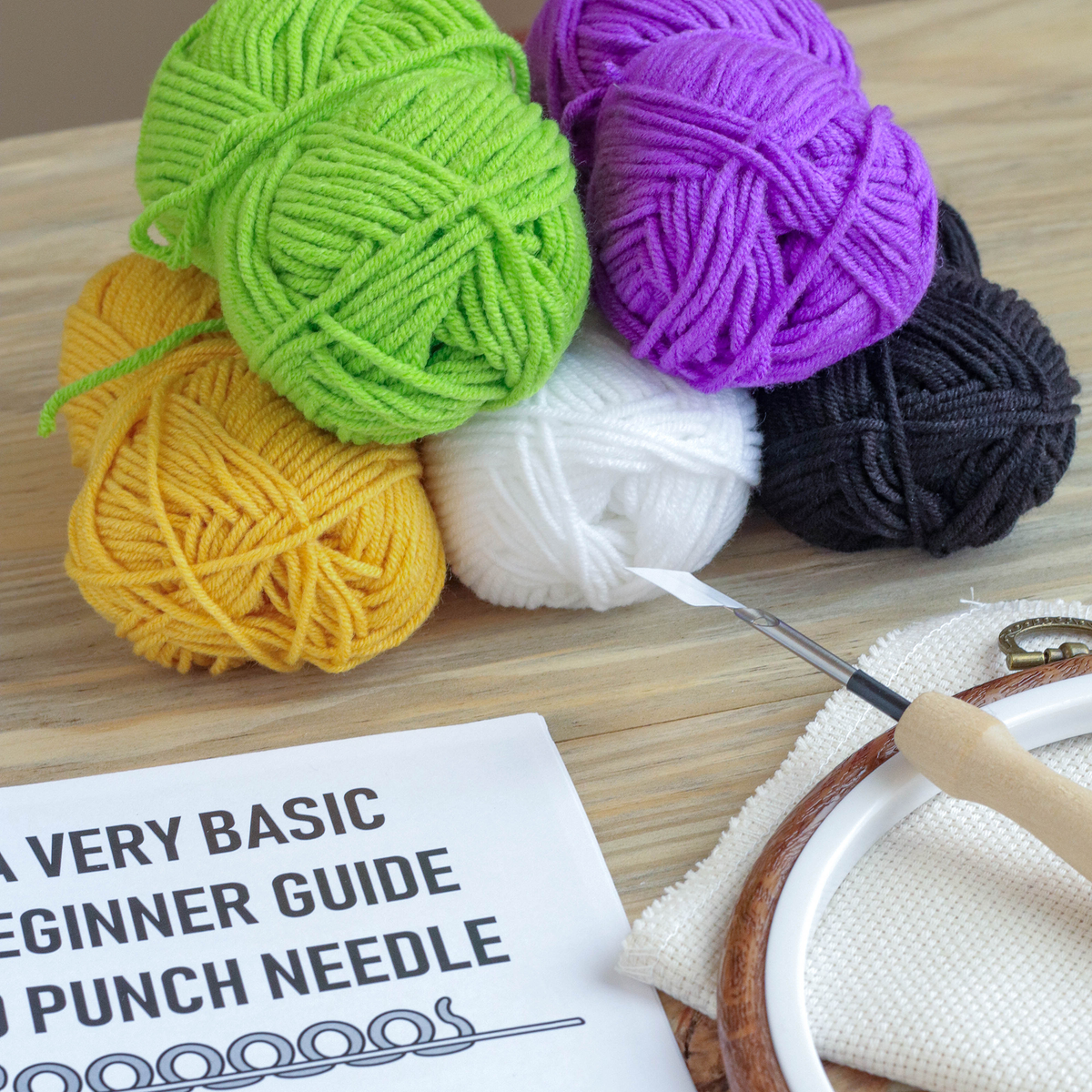 Colorful yarn balls and a punch needle on a wooden surface with a beginner guide.