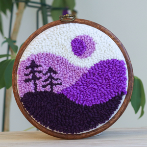 Embroidery hoop with a purple and white landscape design on a wooden surface.
