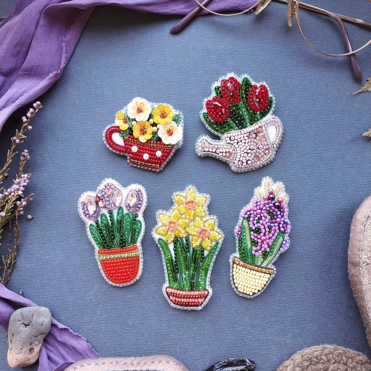 Four colorful beaded flower and plant potted brooches arranged on a textured blue-gray fabric background with decorative purple fabric and stones around the edges.