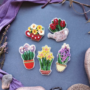 Four colorful beaded flower and plant potted brooches arranged on a textured blue-gray fabric background with decorative purple fabric and stones around the edges.
