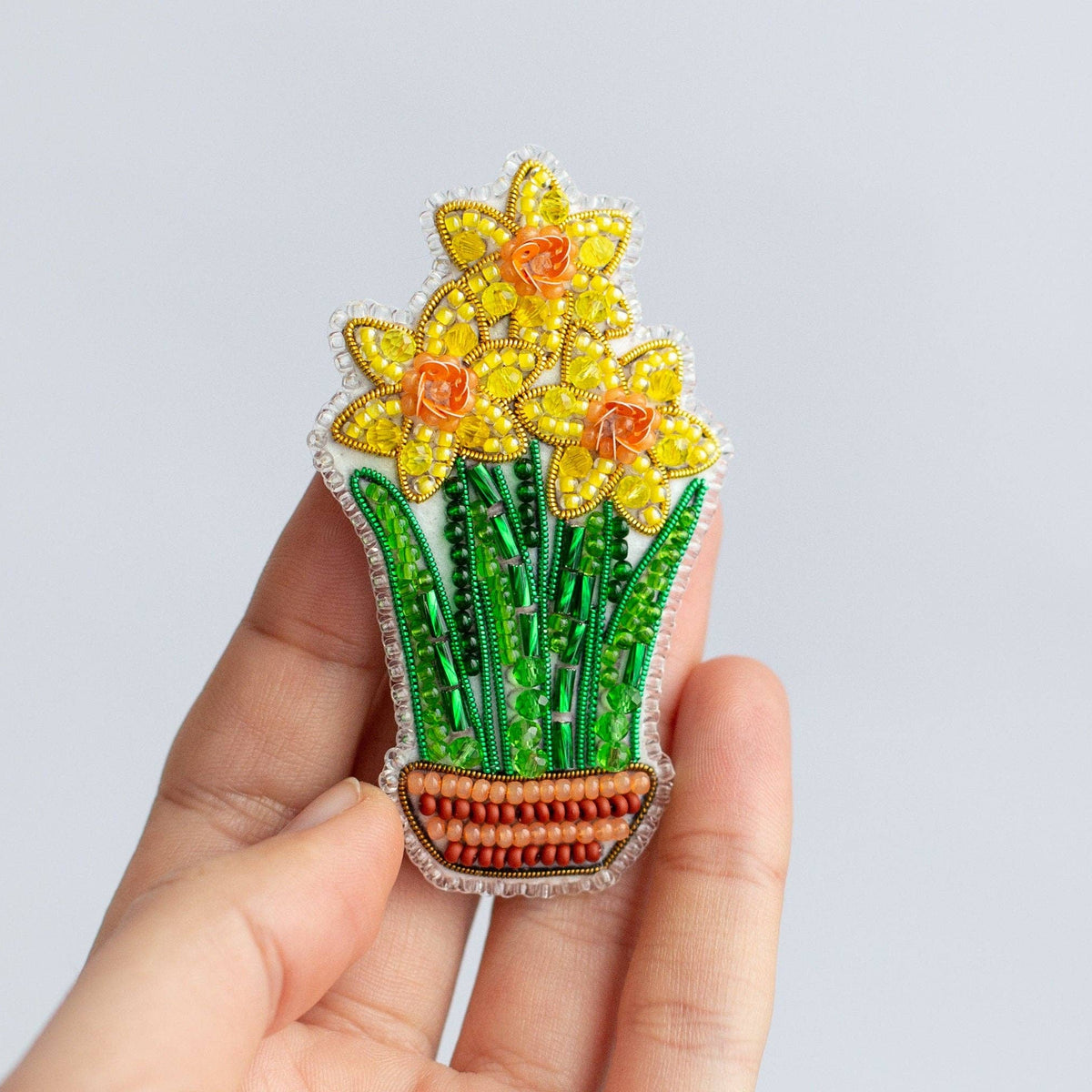 A hand holding a small embroidered brooch of yellow daffodils in a green pot against a plain light gray background.