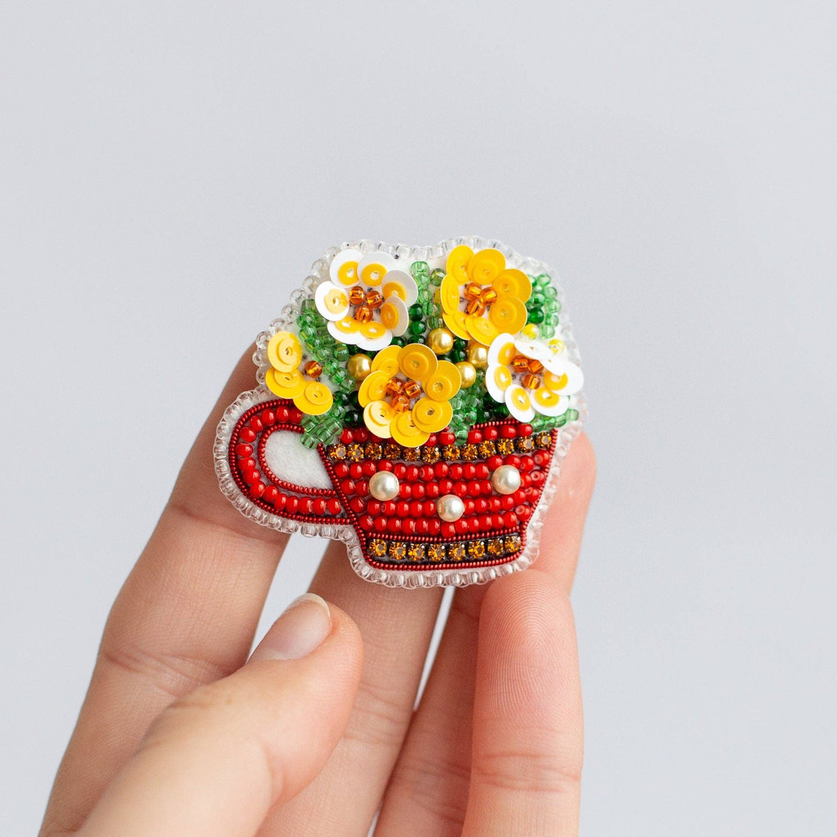 A hand holding a small decorative flower-shaped brooch with a red teacup design, yellow and white flowers, green leaves, and red beads, against a plain light gray background.