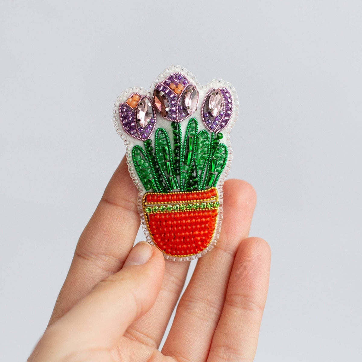 A hand holding a colorful bead embroidered brooch depicting three purple tulips in a red pot with green leaves, set against a plain light gray background.