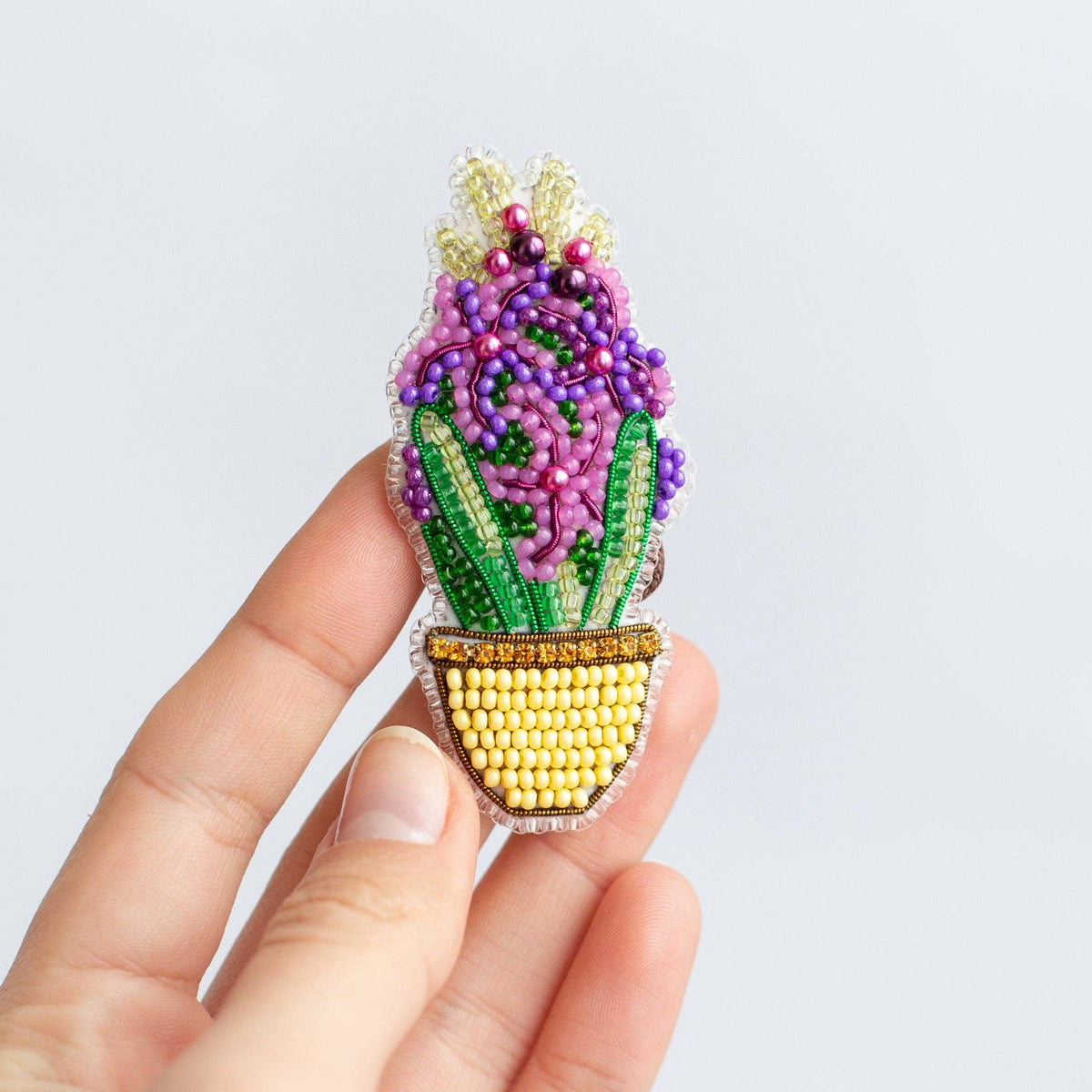 A hand holding a small beaded flower pin in a yellow pot against a plain light gray background.