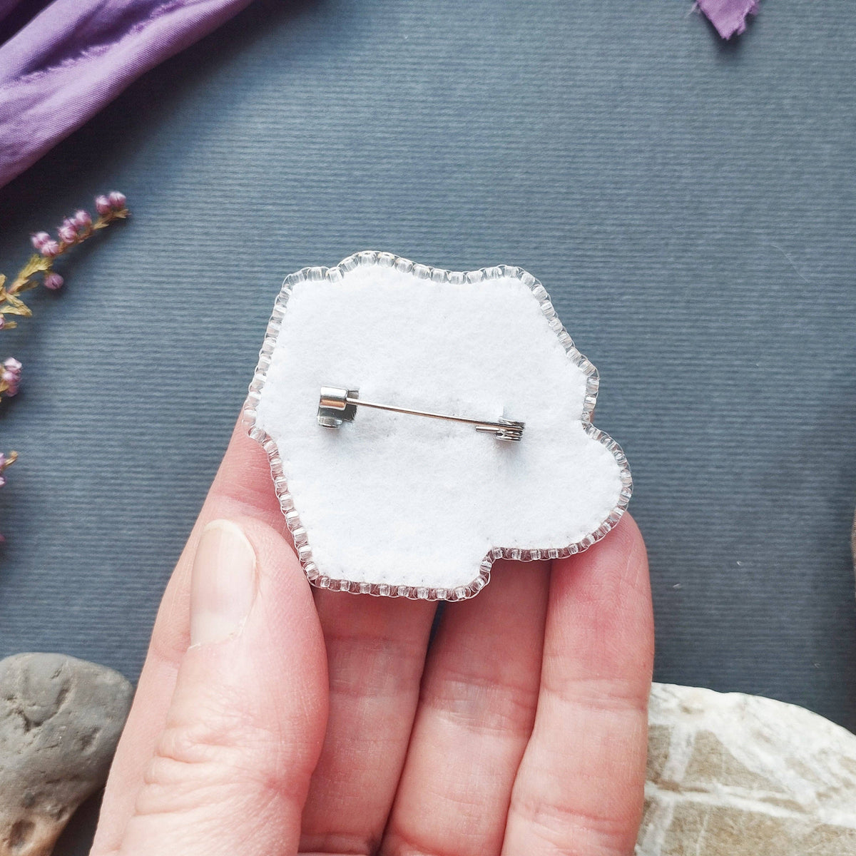 A hand holding a white cloud-shaped brooch with a silver metal border and safety pin back, set against a dark gray background with decorative stones and dried flowers.