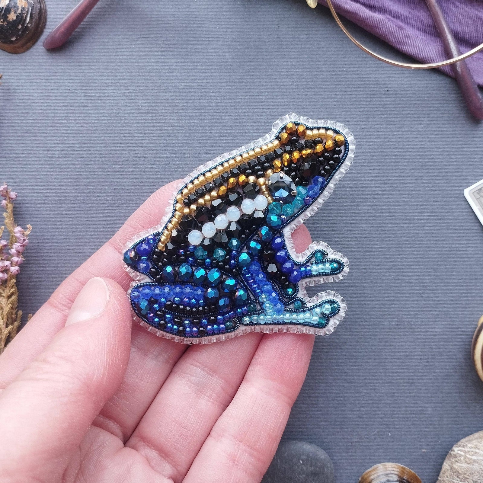 Hand holding a beaded blue frog brooch on a textured surface