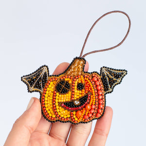 A hand holding a small beaded pumpkin ornament with bat wings and a brown hanging loop, shown against a light gray background.