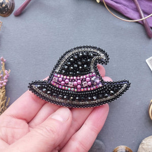 A hand holding a black beaded witch hat brooch decorated with pink and purple rhinestones, set against a gray textured background with mystical props.