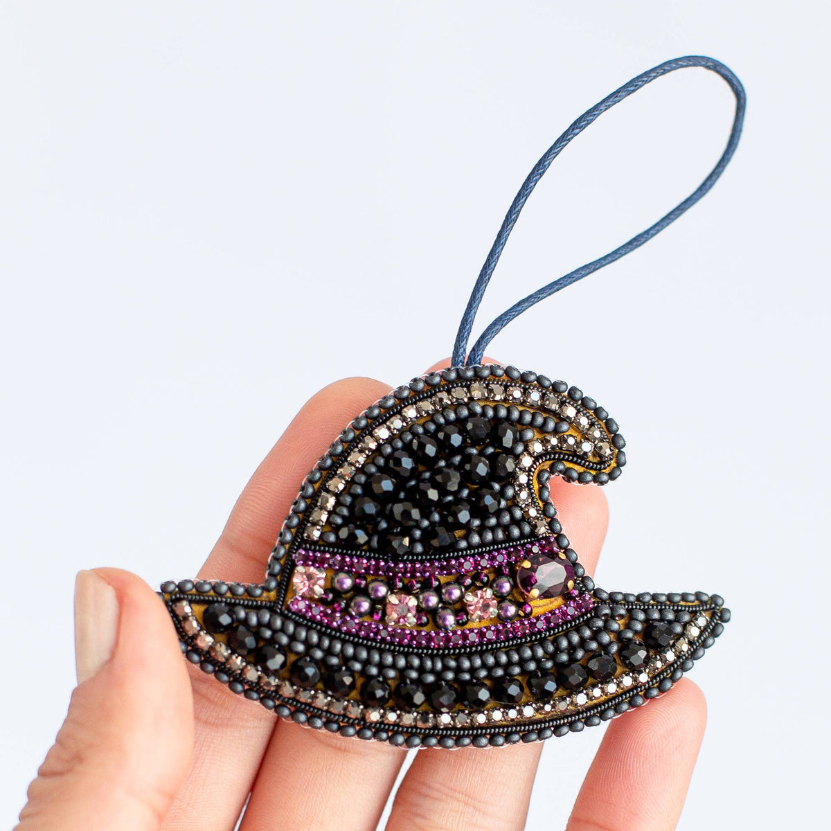 A hand holding a small witch hat ornament made of black seed beads with purple and gold accents, featuring a blue hanging loop, against a plain light gray background.