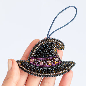 A hand holding a small witch hat ornament made of black seed beads with purple and gold accents, featuring a blue hanging loop, against a plain light gray background.