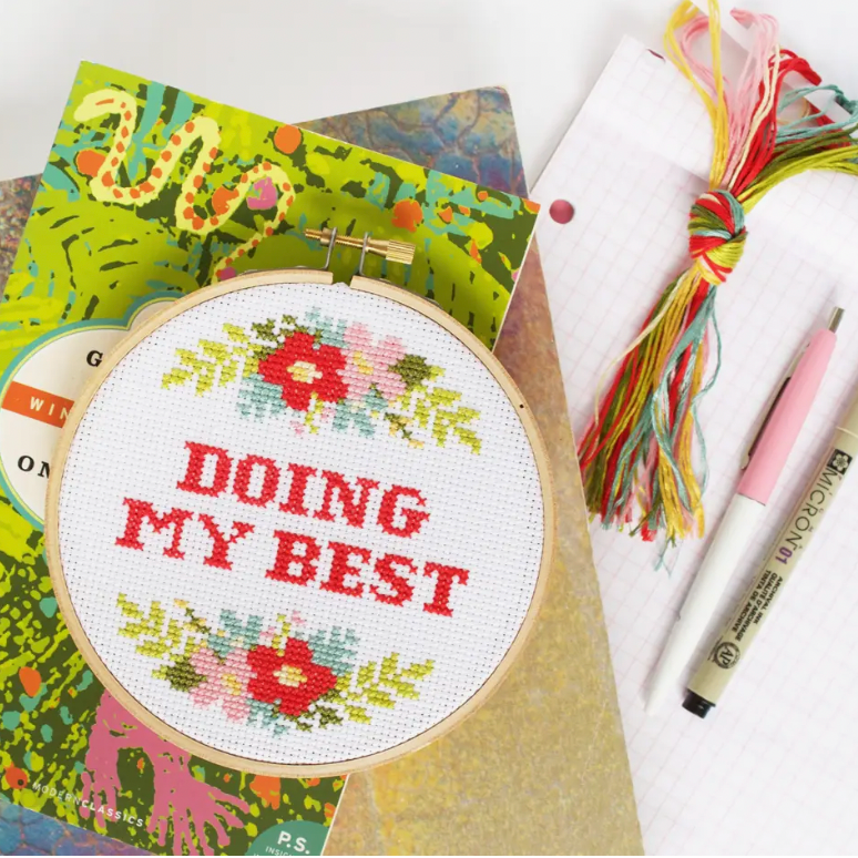 A cross stitch kit with the phrase 'DOING MY BEST' surrounded by a floral pattern, displayed in an embroidery hoop. Embroidery floss and graph paper and some pens are sitting on the right side. 