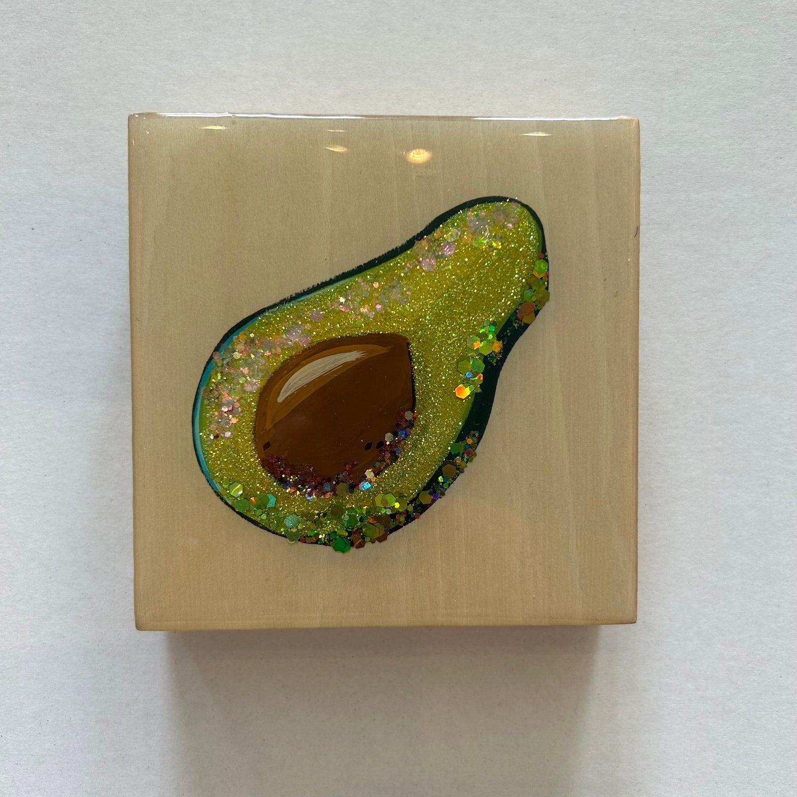 A square beige painting featuring an embedded avocado design with glittery green flesh, a brown seed, and a dark green outer edge, photographed on a plain white background.