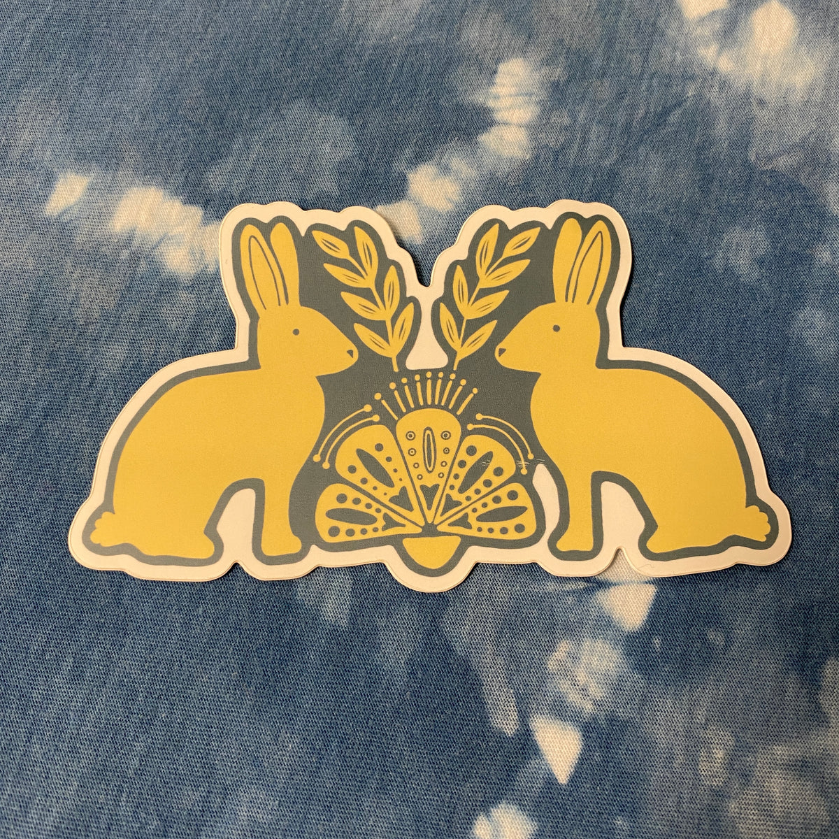 A cream and blut sticker featuring two rabbits facing each other with olive branches and a fan motif in the center, placed on a blue and white tie-dye denim fabric background.