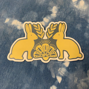 A cream and blut sticker featuring two rabbits facing each other with olive branches and a fan motif in the center, placed on a blue and white tie-dye denim fabric background.