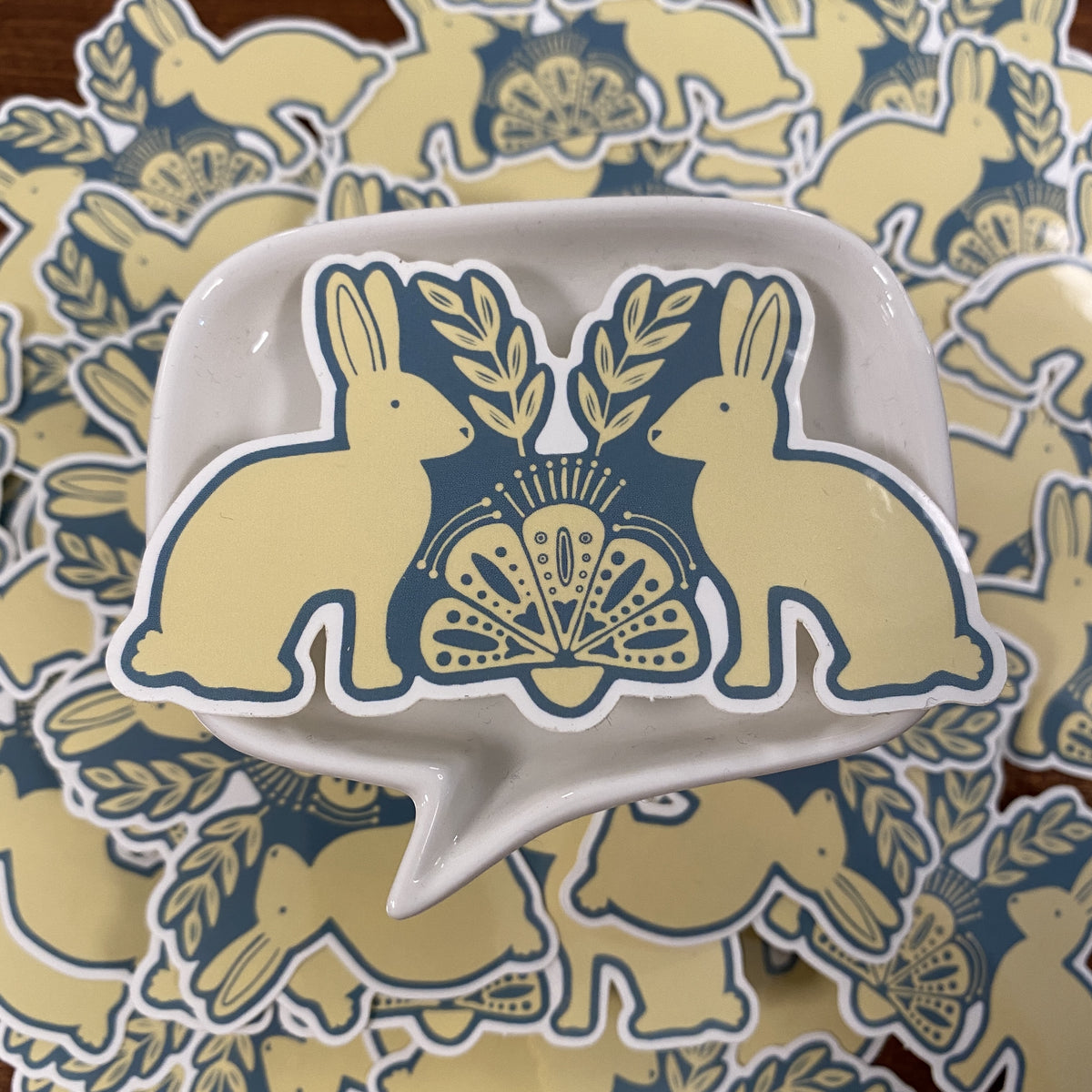 A white ceramic dish with a speech-bubble shape and a blue-and-cream bunny illustration, placed on top of a matching pile of similar stickers.