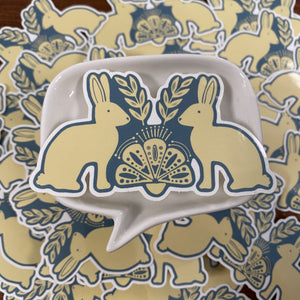 A white ceramic dish with a speech-bubble shape and a blue-and-cream bunny illustration, placed on top of a matching pile of similar stickers.