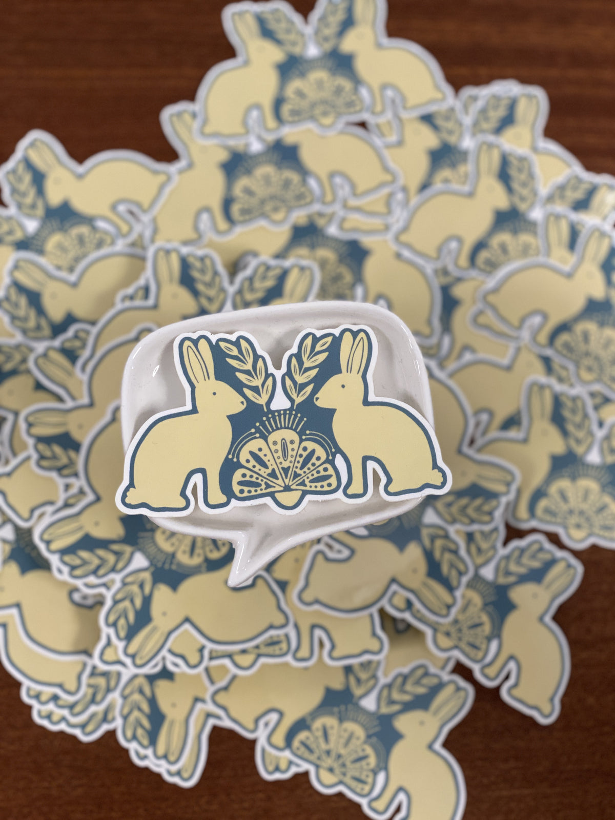A close-up of a white ceramic dish with a blue and pale yellow bunny illustration, surrounded by a pile of matching bunny-patterned stickers. 
