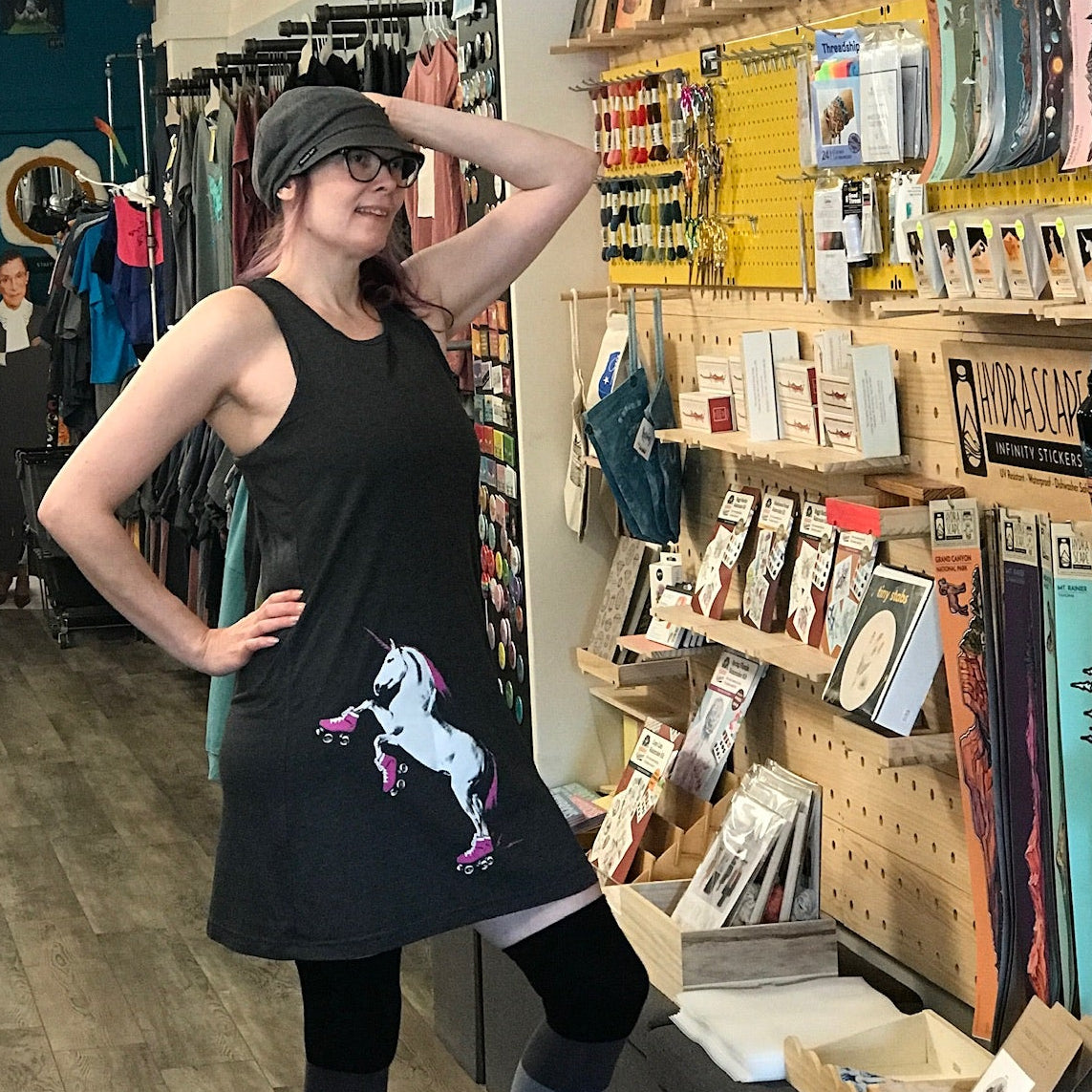 Woman in a store with shelves and posters on the wall. She is wearing a black tank dress with a roller skating unicorn on the skirt. 
