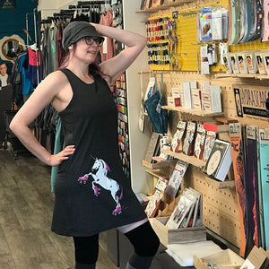 Woman in a store with shelves and posters on the wall. She is wearing a black tank dress with a roller skating unicorn on the skirt. 