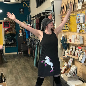 Person in a store with arms outstretched, surrounded by shelves with books and items. They are wearing a black tank dress with a roller skating unicorn on the skirt.