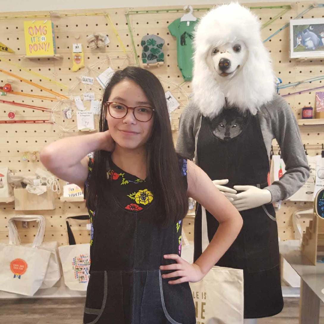 Woman posing with a mannequin wearing a black apron dress in a store setting.