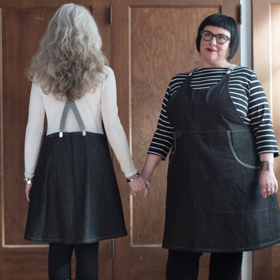 Two women wearing black aprons holding hands in front of a wooden door.