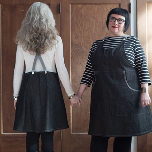 Two women wearing black aprons holding hands in front of a wooden door.