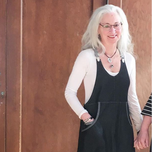 Woman with gray hair wearing glasses and a black apron dress standing in front of a wooden door.