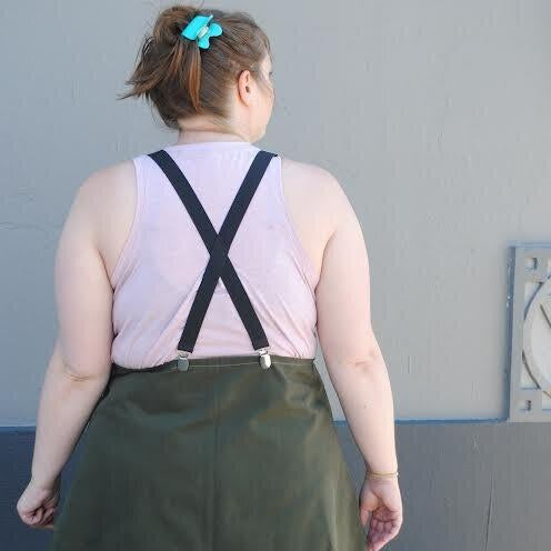 Person wearing a pink sleeveless top and green skirt with suspenders against a gray wall.