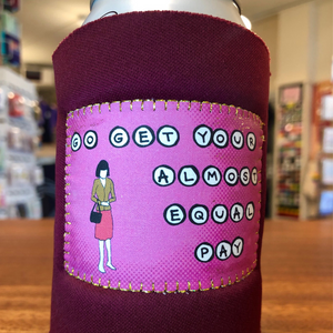 A close-up of a beverage can wrapped in a burgundy fabric can cooler sleeve with a pink patch that reads 'GO GET YOUR ALMOST EQUAL PAY' with a small illustrated woman holding a bag. The background shows a blurred retail store interior and wooden tabletop.