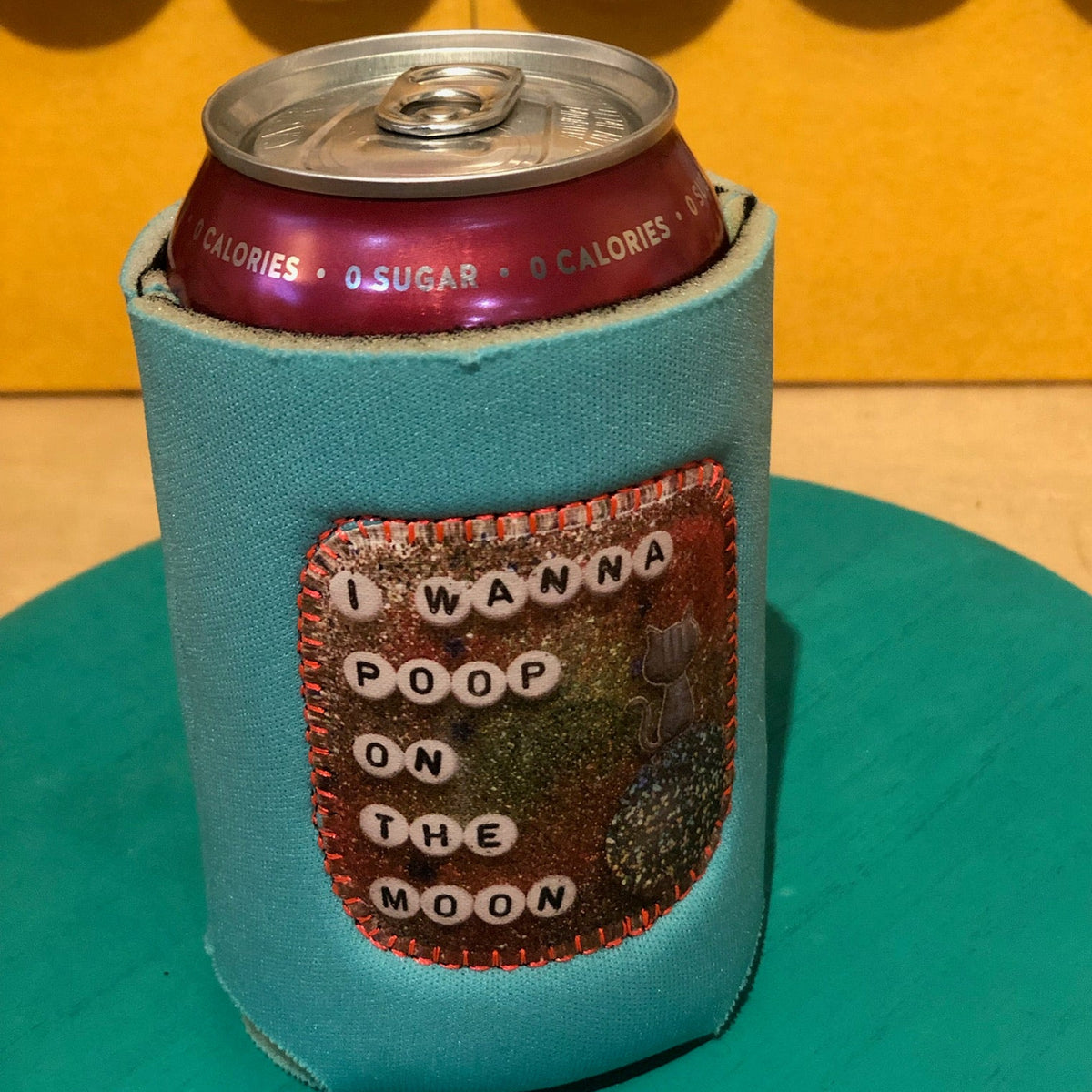 A turquoise neoprene can cooler sleeve with a glittery patch reading 'I WANNA POOP ON THE MOON' in white letter tiles, holding a dark red beverage can, set against a yellow background.