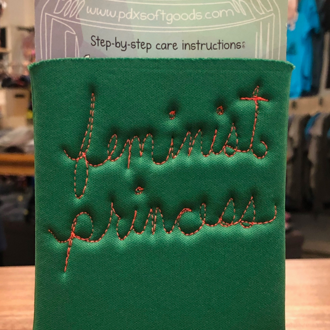 A green fabric can cooler sleeve with 'feminist princess' stitched in orange thread.