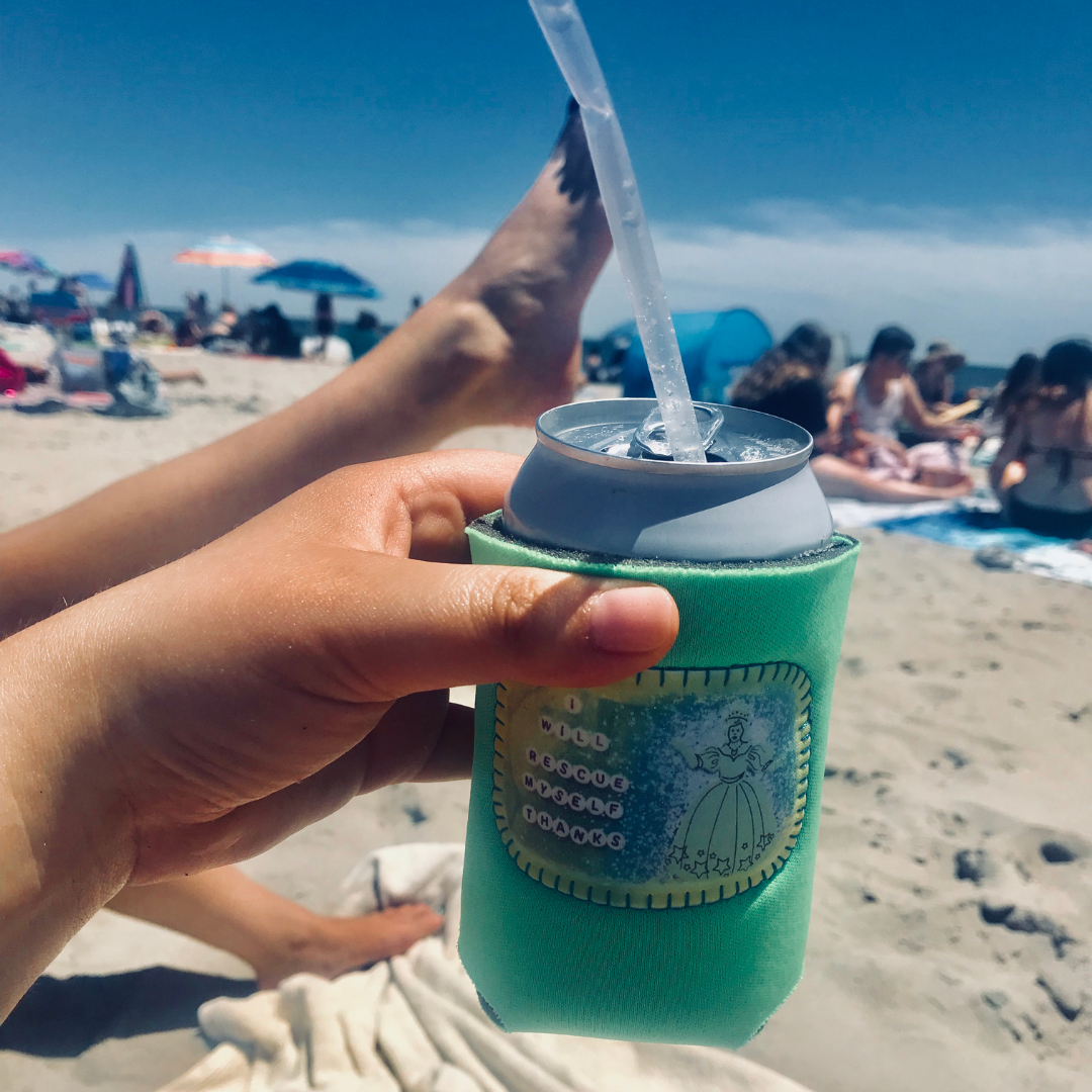A hand holds a green neoprene can cooler with a clear straw and a silver can inside, photographed on a sunny beach with sand, people, and umbrellas in the background.