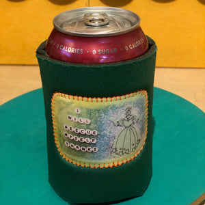A dark green neoprene can cooler sleeve with a colorful embroidered patch featuring a cartoon character and the text 'I WILL RESCUE MYSELF, THANKS' holding a red can on a teal surface.