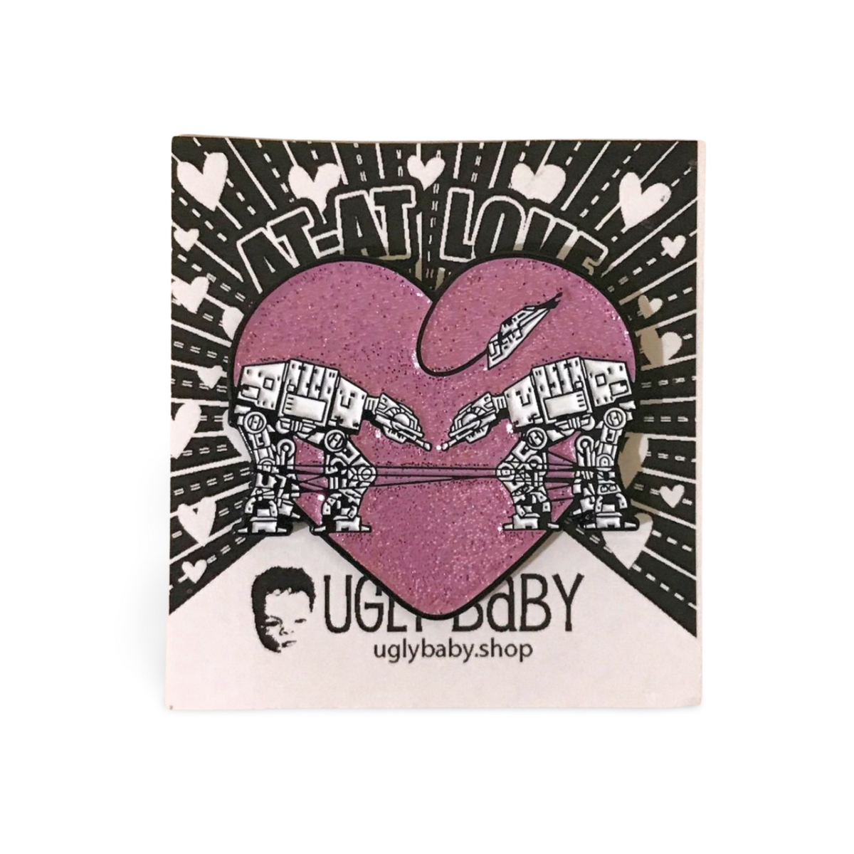 Heart shaped enamel pin called Love AT-AT First Sight with a pink glitter background. Shown on a backing card with hearts. 