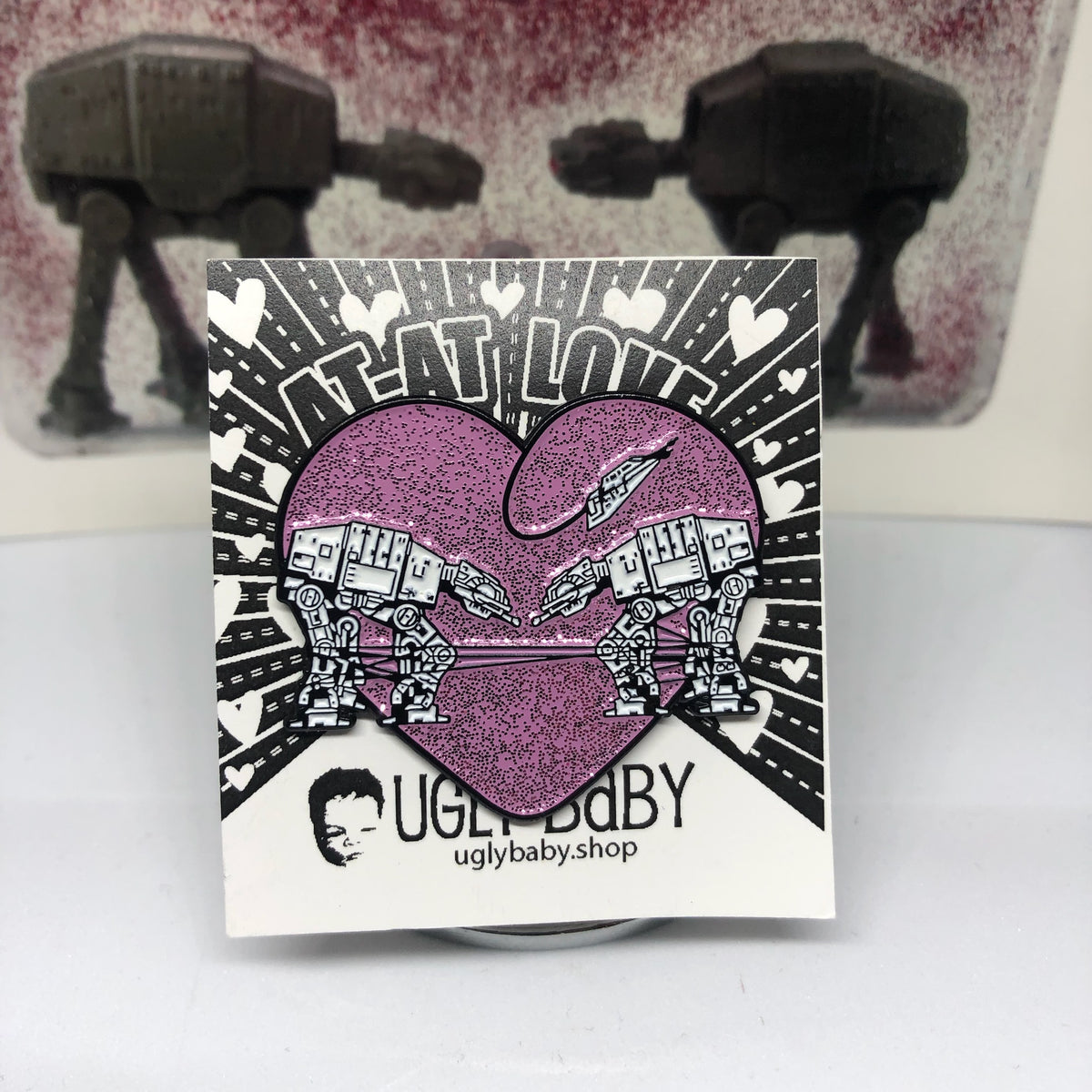 Heart shaped enamel pin called Love AT-AT First Sight with a pink glitter background. Shown on a backing card with hearts. 