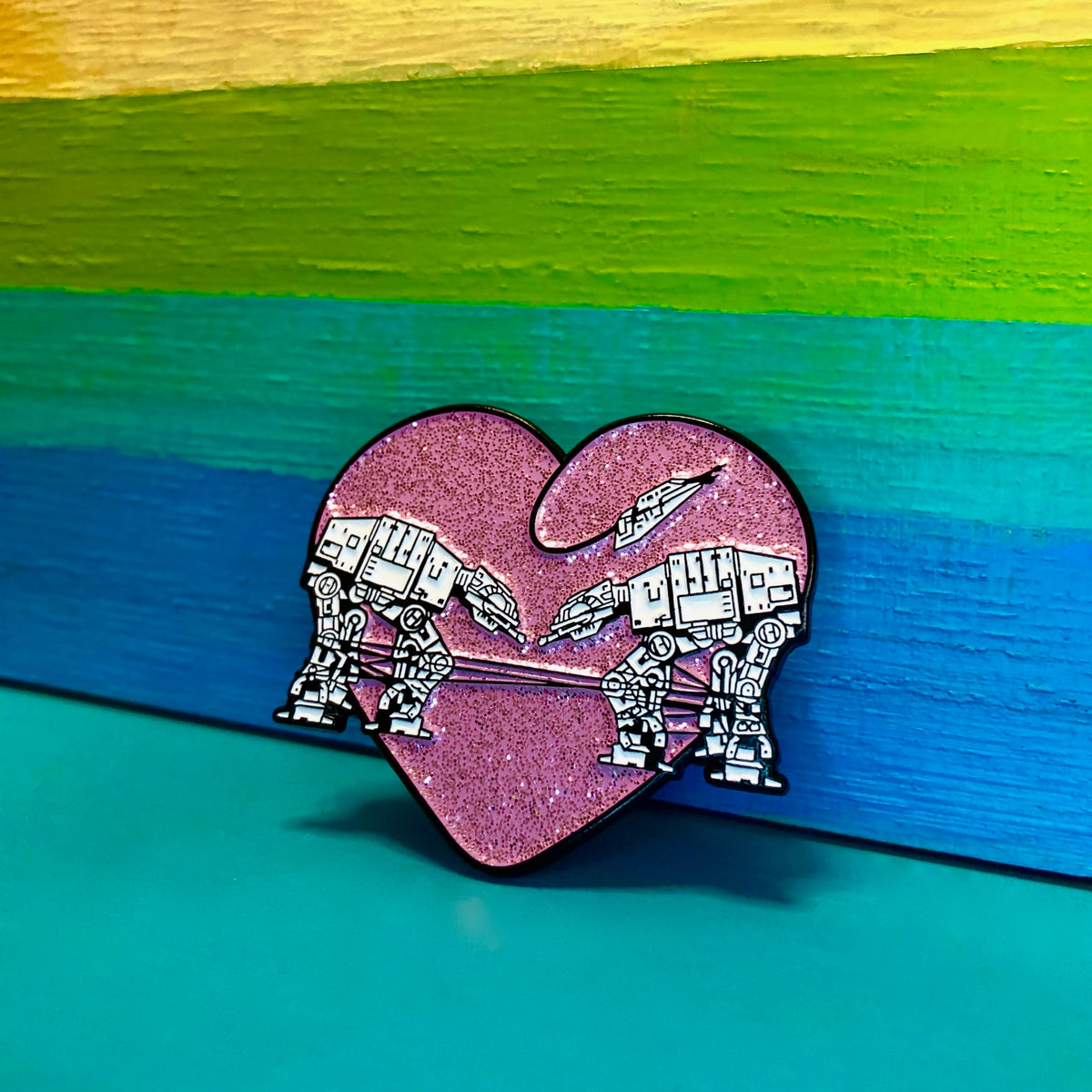 Heart shaped enamel pin called Love AT-AT First Sight with a pink glitter background. Shown against a rainbow background. 